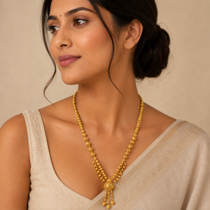 Woman wearing a 22K gold traditional beaded tassel necklace featuring intricately textured gold beads, cascading bead clusters, and a decorative tassel pendant, paired with a beige saree for an elegant ethnic look.
