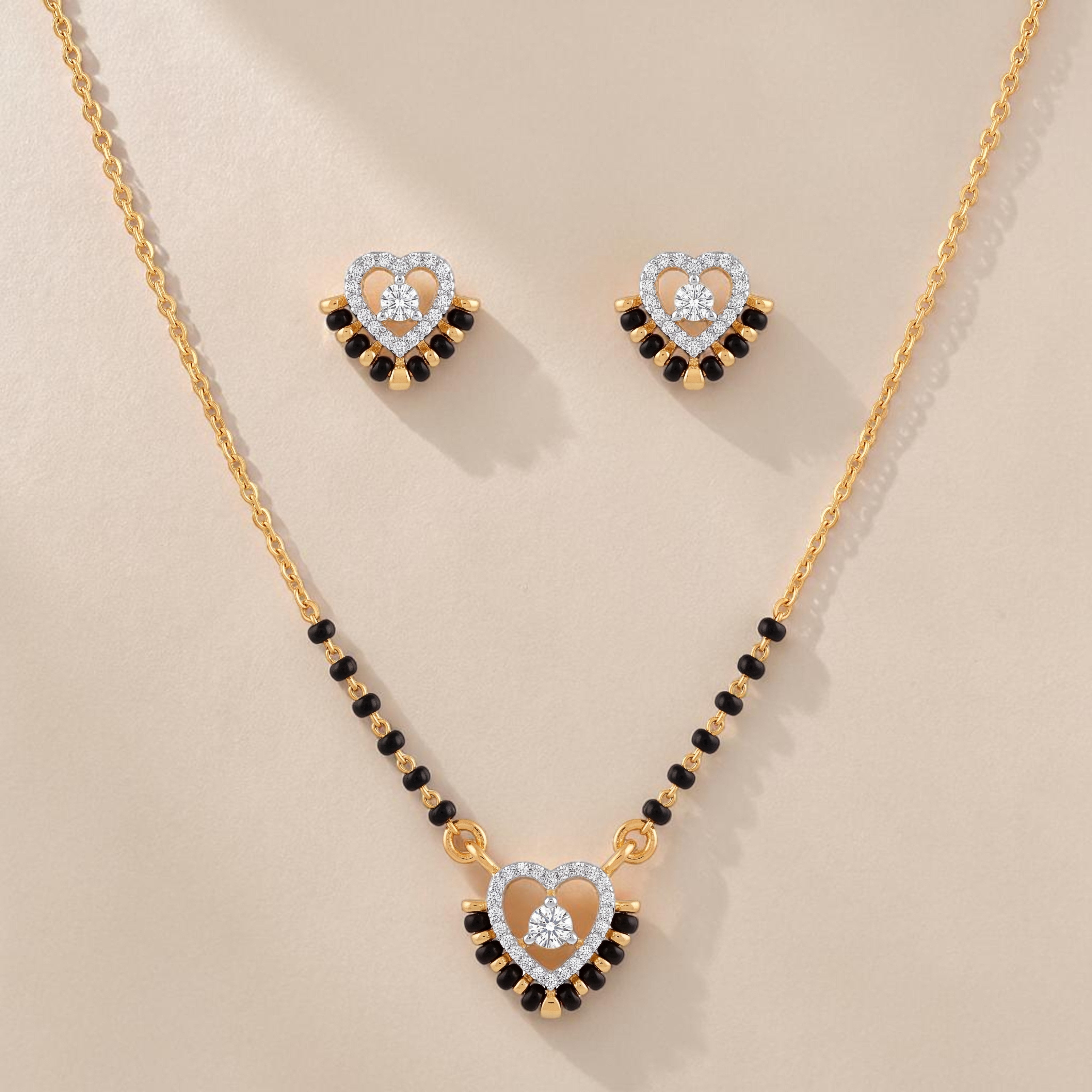 Elegant gold mangalsutra jewelry set featuring black beads, a heart-shaped diamond pendant, and matching heart stud earrings on a white background.