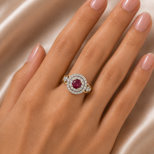 Elegant gold ring featuring a vivid oval ruby center surrounded by a sparkling double diamond halo, with additional diamond accents along the shoulders.