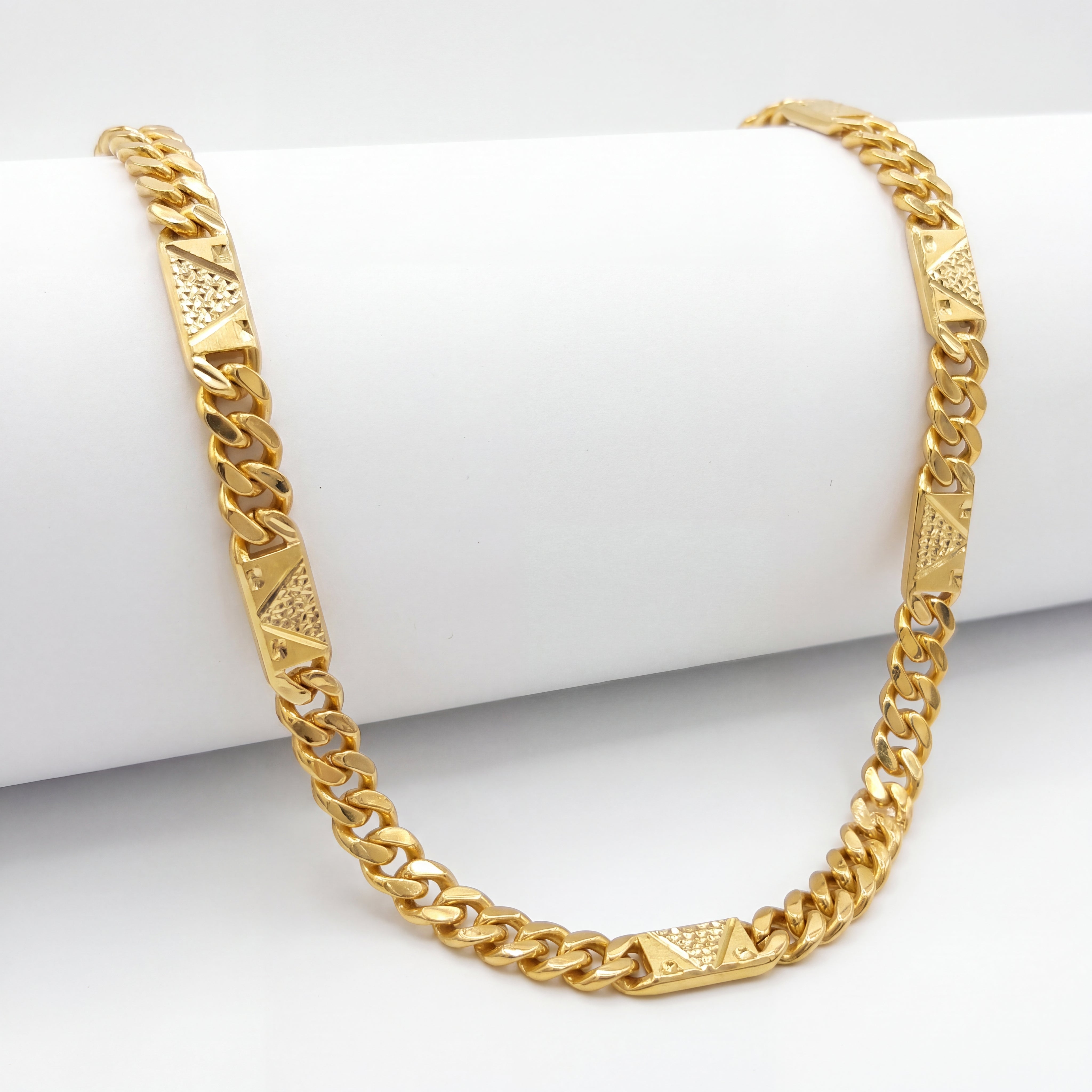 Bold 22K yellow gold Cuban link chain necklace featuring geometric textured designer plates and polished interlocking links displayed on a white cylindrical stand.