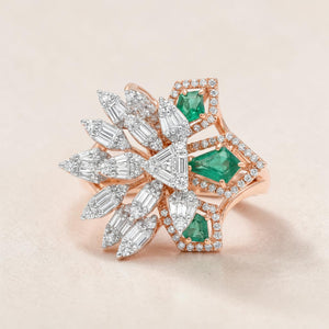 Rose gold statement ring featuring asymmetrical design with vibrant emerald-cut green gemstones, surrounded by baguette and marquise diamonds arranged in a floral-inspired burst with pavé diamond accents.