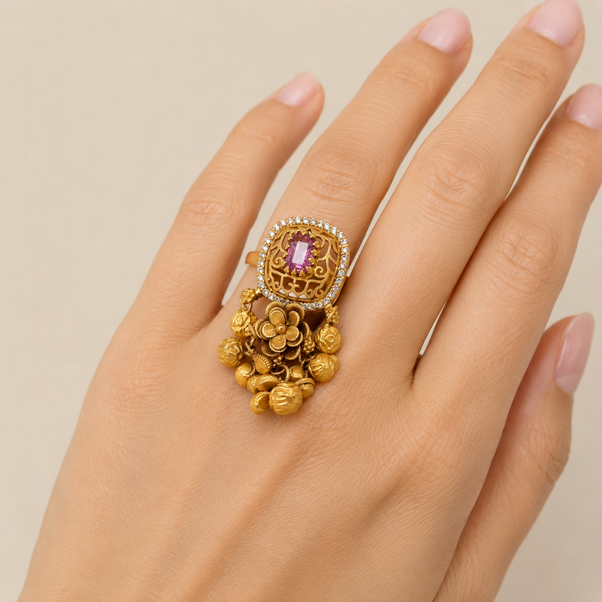 Traditional gold statement ring featuring a rectangular purple-pink gemstone framed by intricate filigree work and diamond accents, finished with ornate floral detailing and multiple dangling gold bead charms.