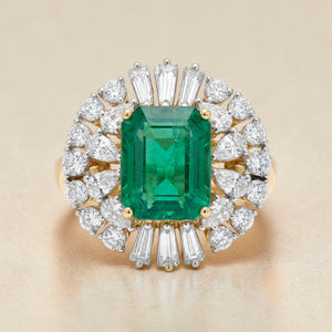 Yellow gold ring featuring a striking emerald-cut green gemstone center, surrounded by a radiant halo of pear-shaped and baguette diamonds arranged in a burst design for a bold and luxurious look.