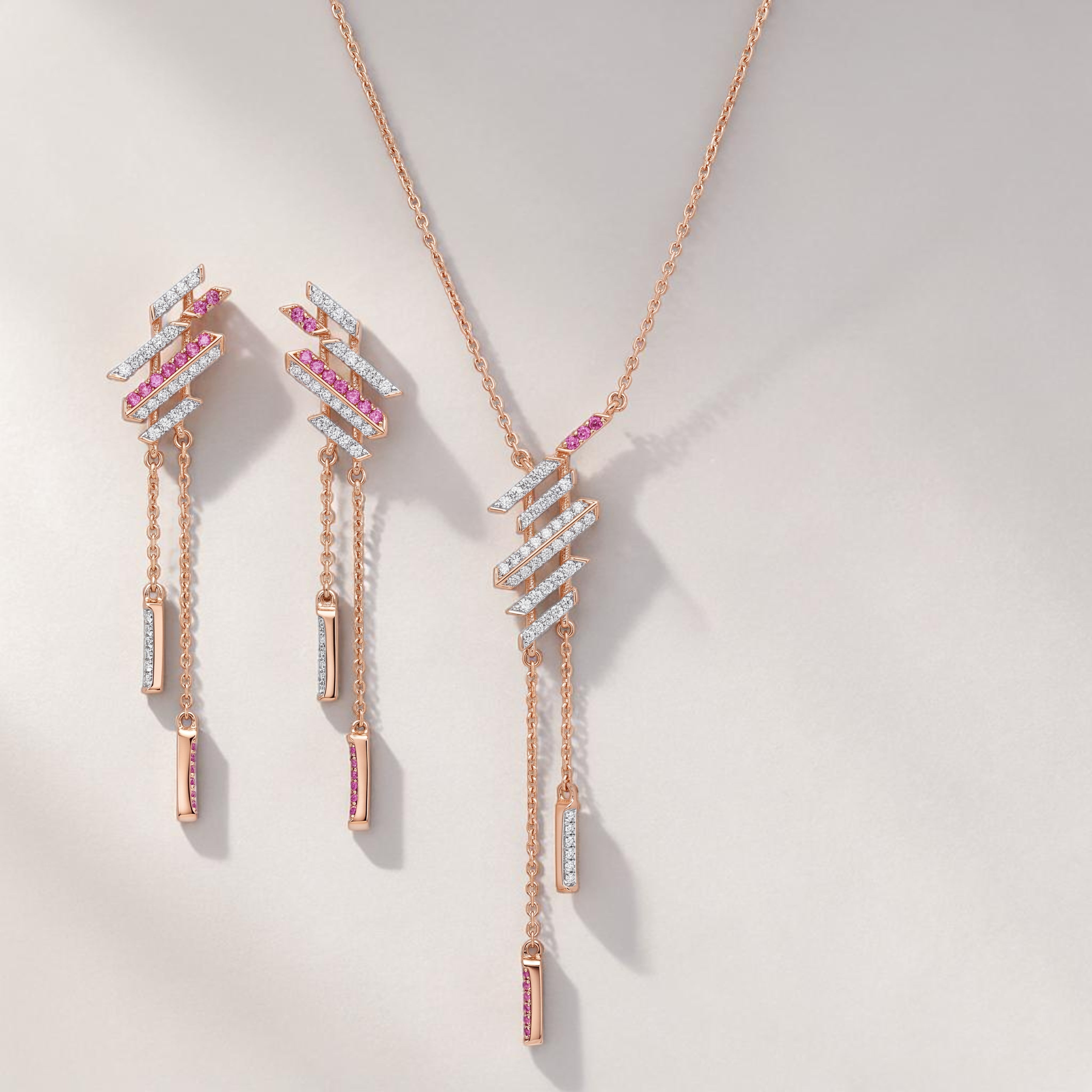 Modern rose gold jewelry set featuring geometric diamond and pink sapphire accents with dangling bar details in a pendant necklace and matching earrings on a white background.
